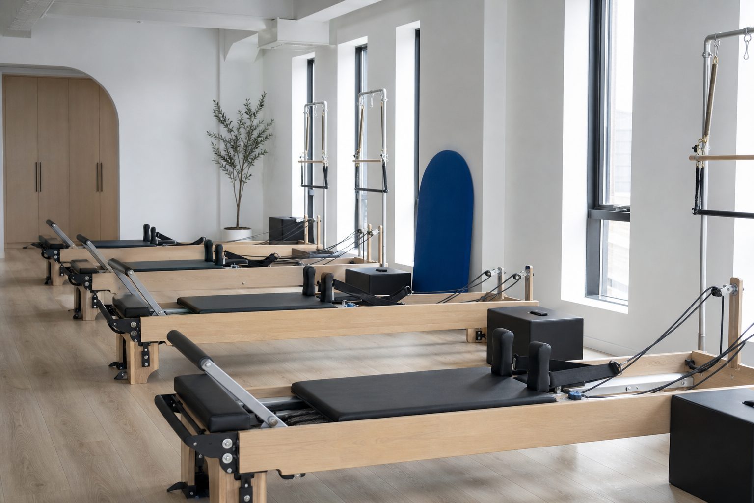 Row of Pilates reformers spaced evenly across a bright chalk white studio floor lit by tall windows