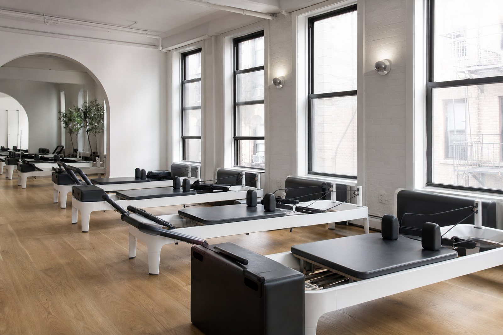An empty Pilates studio with a row of reformers on a wooden floor lit by tall windows